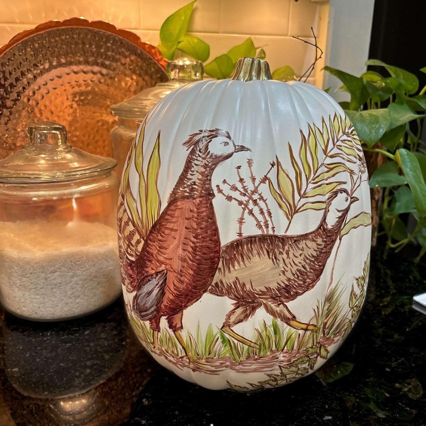 Hand-painted ivory decorative pumpkin featuring two pheasants in rich fall colors walking through foliage, with warm ambient lighting on a kitchen counter.