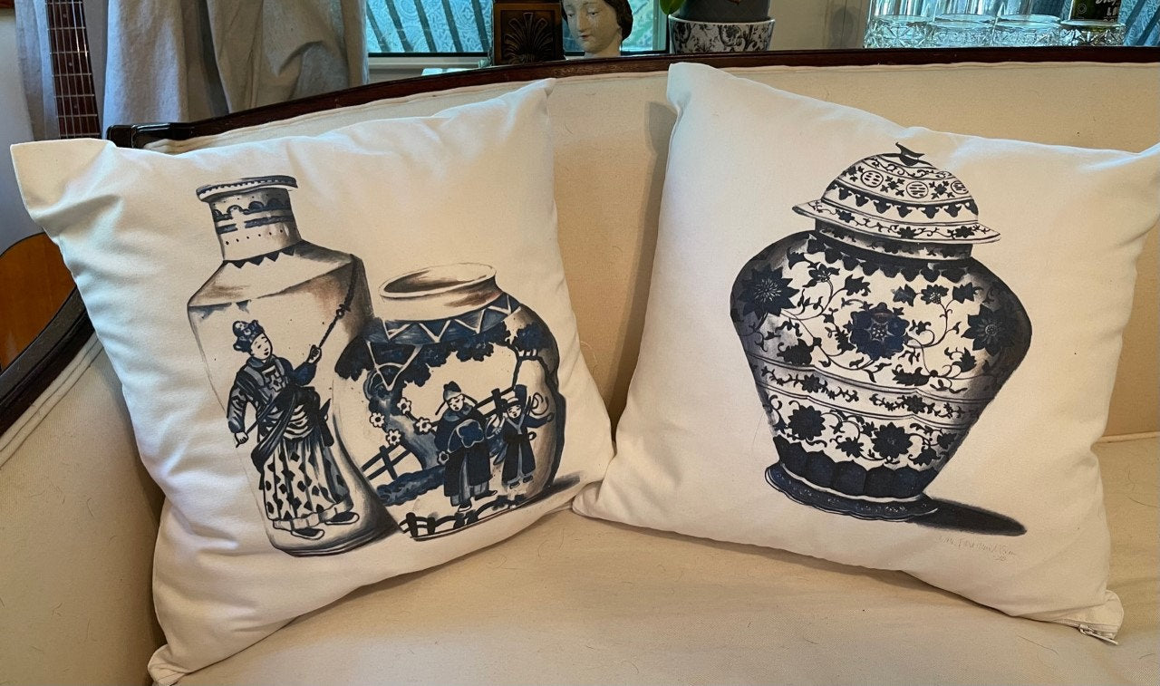 Photo of a two square pillows on a couch. One has a blue chinoiserie floral vase design and the other has two chinoiserie vases with depictions of Chinese people in traditional garments.