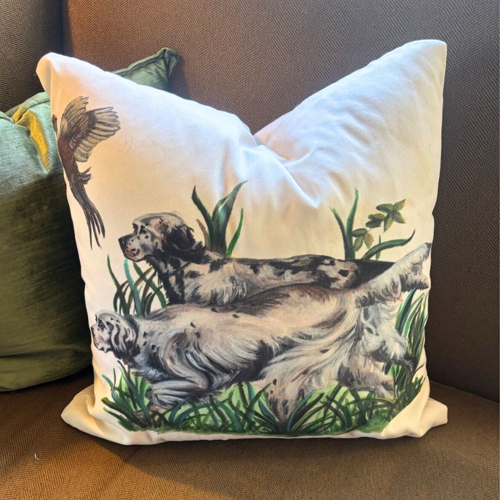 Decorative pillow featuring two English setter bird dogs pointing in tall grass with a bird in flight; rustic-style design on neutral fabric, photographed on a brown couch.