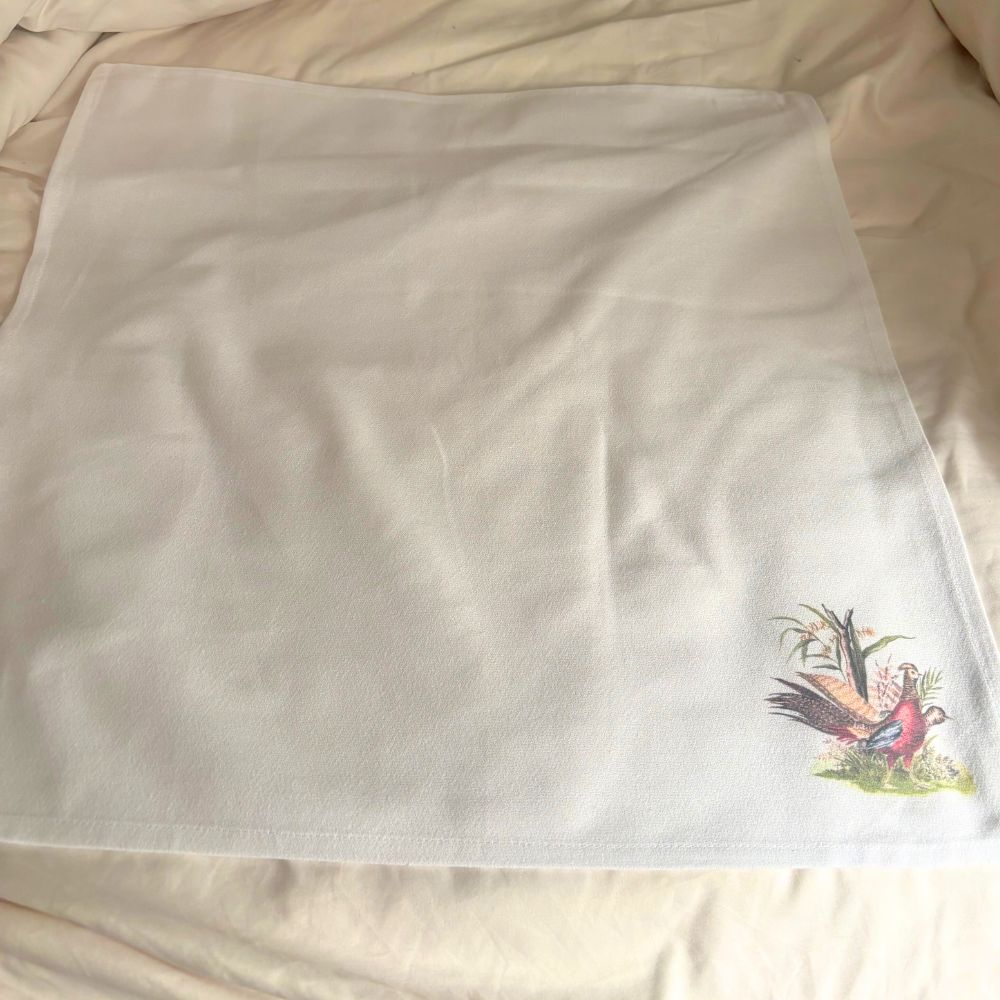 Unfolded cloth napkin with a vintage-style illustration of two pheasants and corn stalks in the bottom right corner.