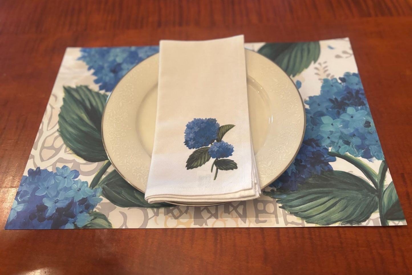 Placemat with a blue hydrangea floral design, plate, and folded white napkin with a single blue hydrangea design on a wooden table