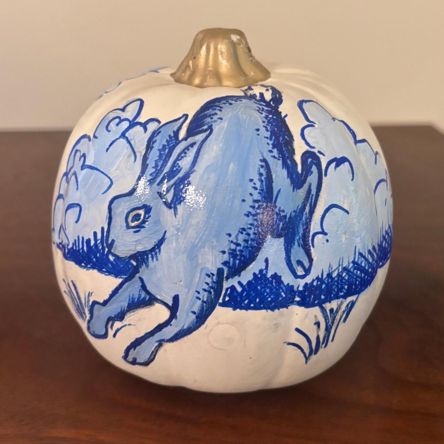 A decorative hand-painted white pumpkin, featuring a blue rabbit in motion with stylized clouds surrounding it. The pumpkin maintains the chinoiserie-inspired aesthetic with rich blue detailing and a metallic gold stem, resting on a wooden surface.