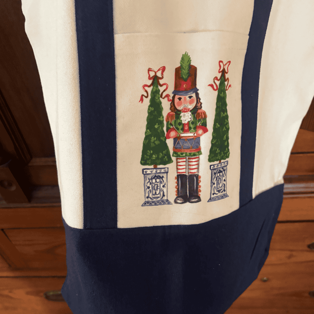 Canvas tote bag with hand-painted nutcracker design, flanked by two holiday topiaries in blue chinoiserie planters