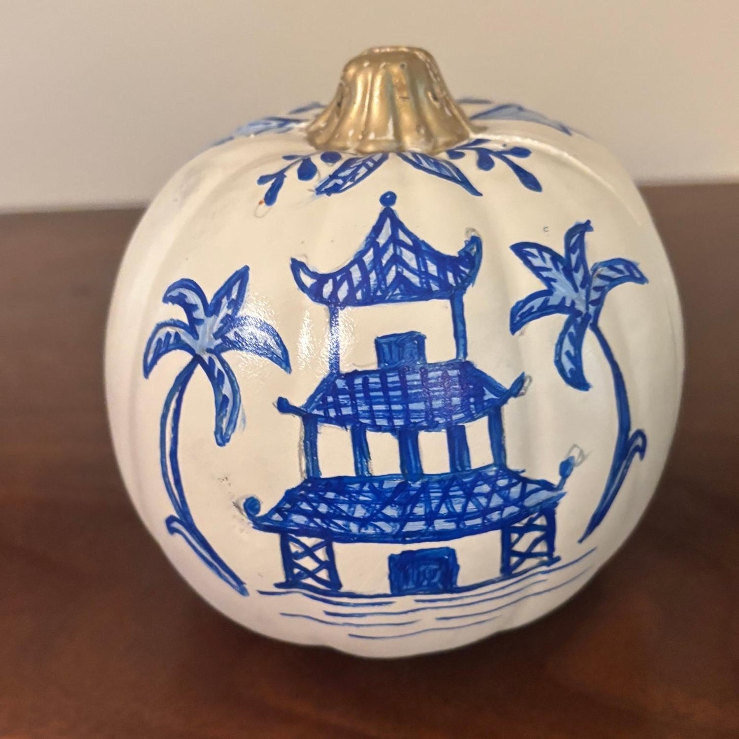 Hand-painted white pumpkin featuring a blue chinoiserie temple design with palm trees and a gold stem.