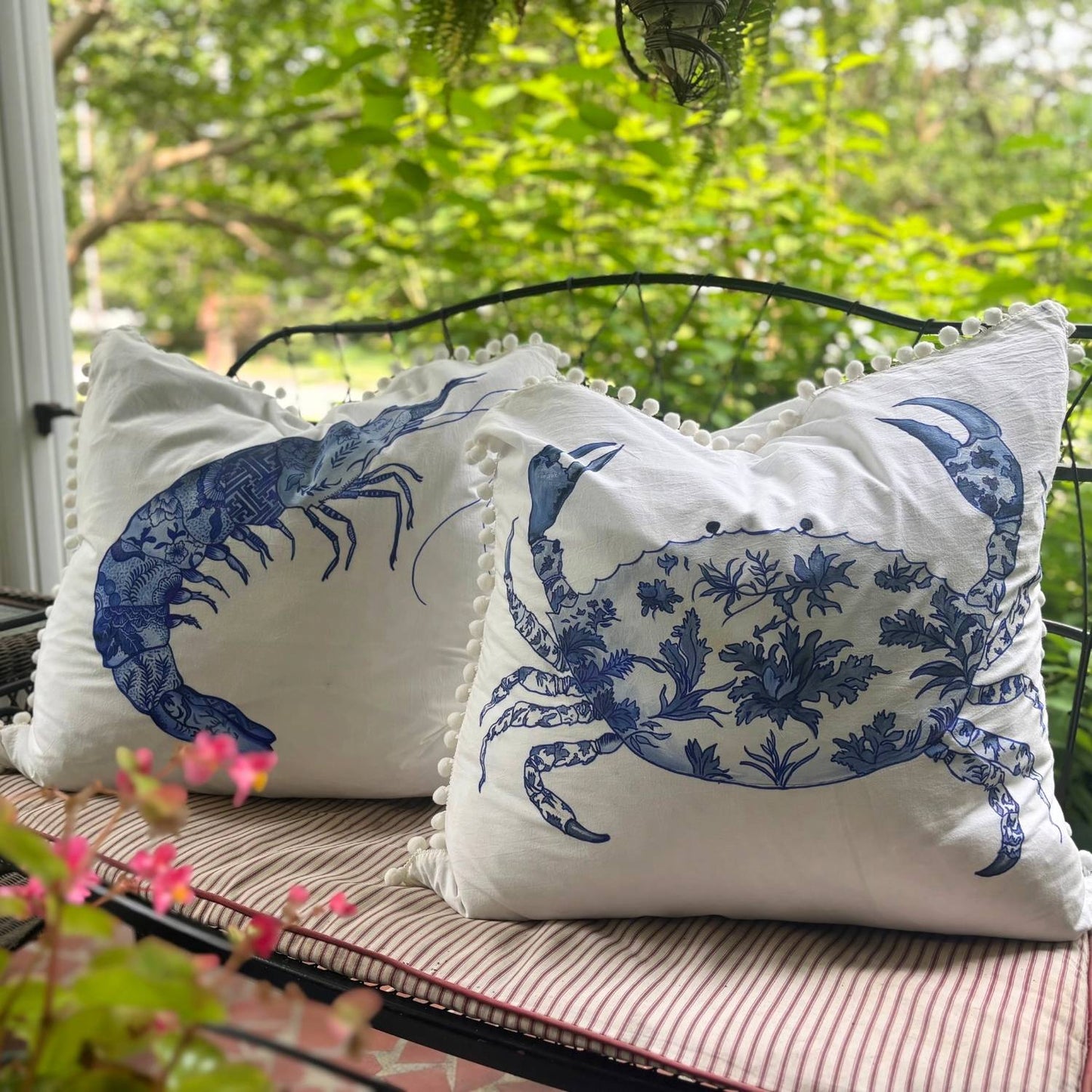 Two hand-painted chinoiserie-style crab and prawn pillows with white pom pom trim, styled on a bench.