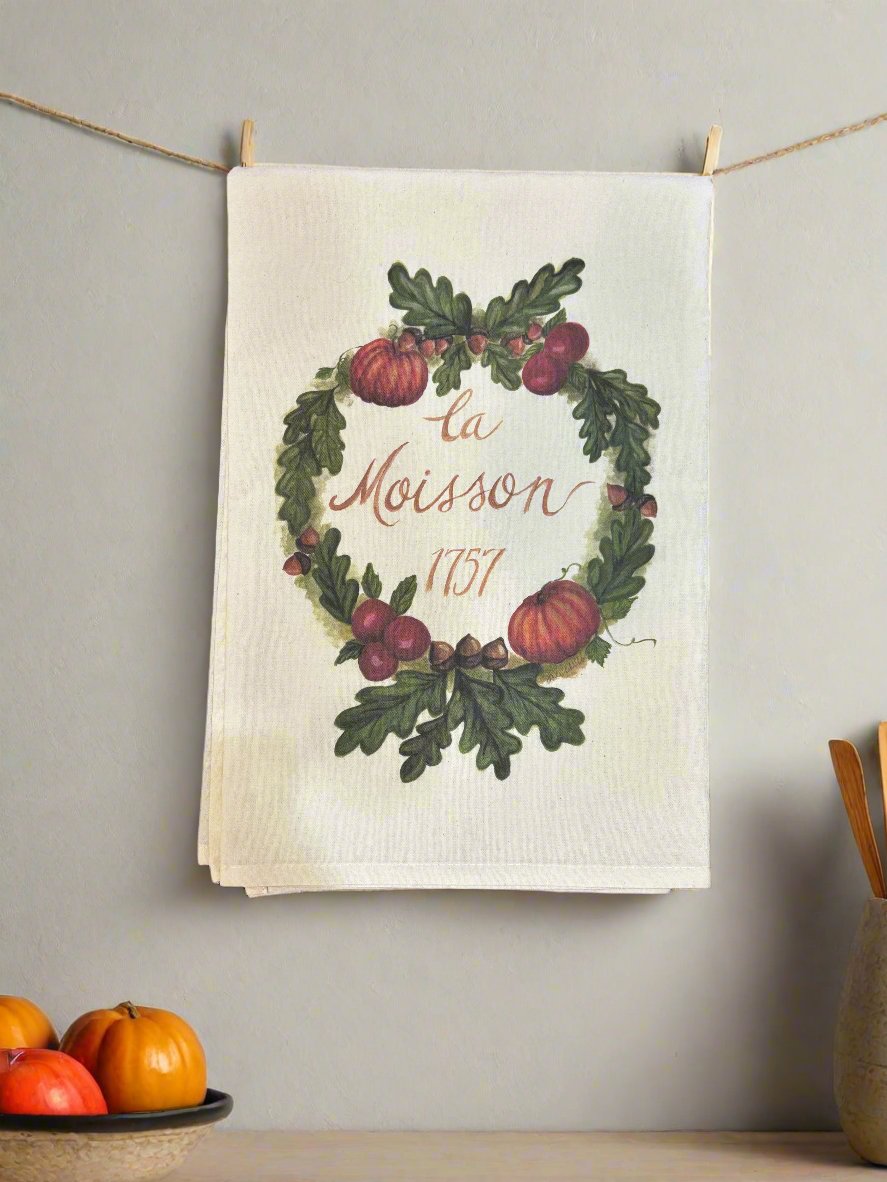 Photo of a white cotton tea towel with a fall harvest wreath design featuring pumpkins, cranberries, acorns, and green oak leaves. It is hanging on twine with clothes pins on a kitchen wall, and small decorative pumpkins are in a bowl on the countertop.