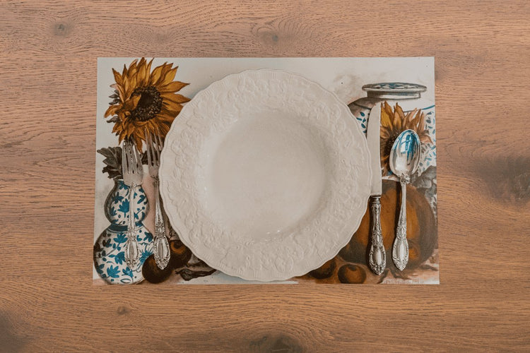 Overhead photo of a fall harvest placemat featuring a vase with sunflowers, pumpkins, and apples with a cream colored plate and silverware on a wooden table