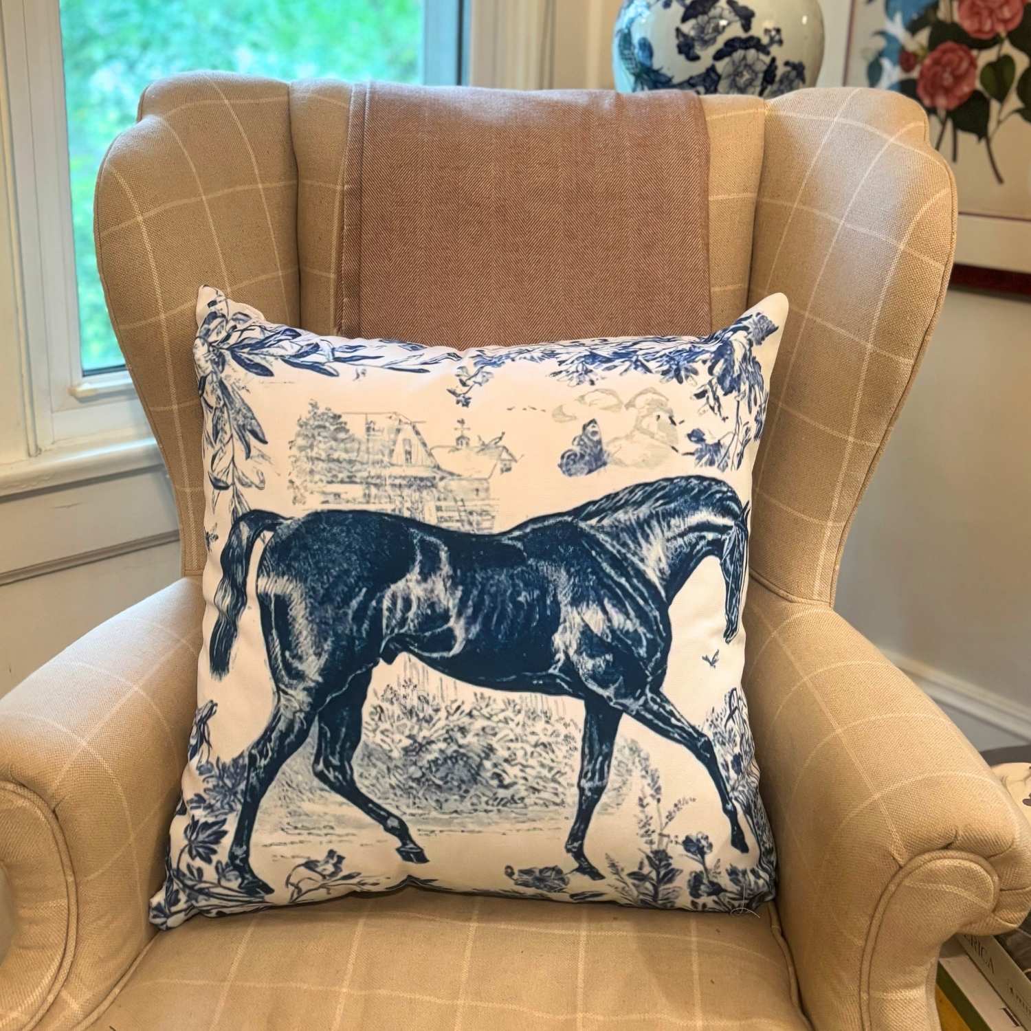 Blue and white decorative throw pillow featuring a detailed navy illustration of a thoroughbred horse on a toile-style background, placed on a beige plaid armchair.