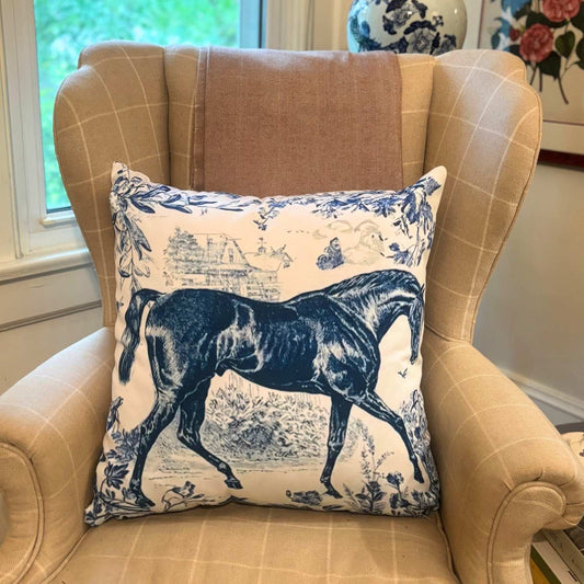 Blue and white decorative throw pillow featuring a detailed navy illustration of a thoroughbred horse on a toile-style background, placed on a beige plaid armchair.