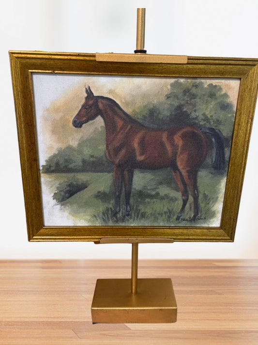 Framed tabletop art on a brass stand featuring a chestnut horse in a soft countryside landscape with green trees and grass.
