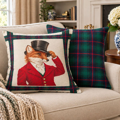 Two decorative throw pillows on a beige sofa in a living room setting. The front pillow features a fox dressed in a red coat and black top hat on a cream background with a green tartan border, while the rear pillow is solid green tartan plaid.