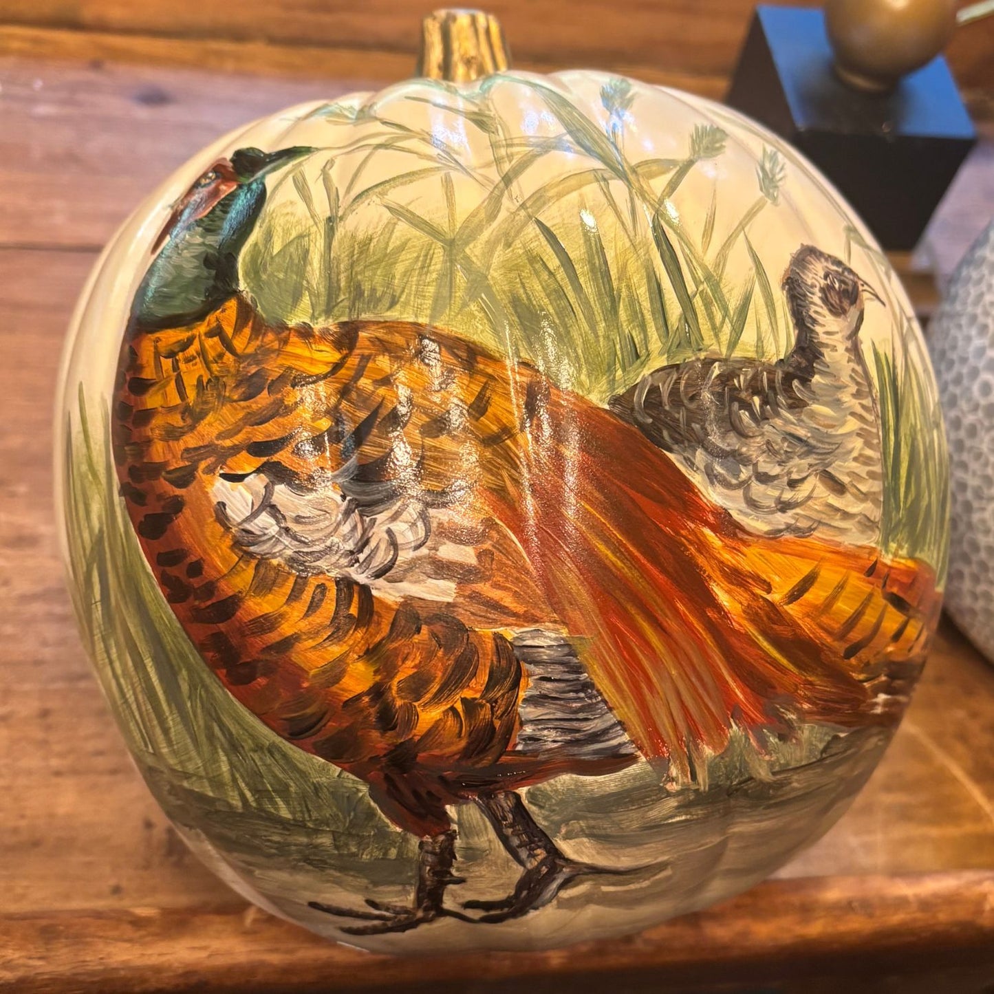 Decorative pumpkin with hand-painted pheasant pair among soft green grass, golden stem accent, and autumnal tones.