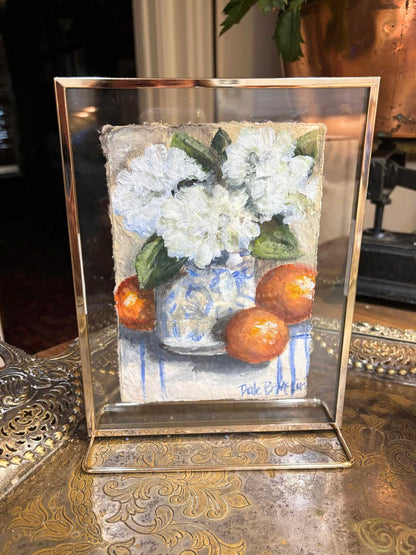 Original acrylic still life painting of white flowers and orange citrus in a blue and white porcelain vase on handmade Lokta paper, framed in a brass floating frame with clear glass for tabletop display.