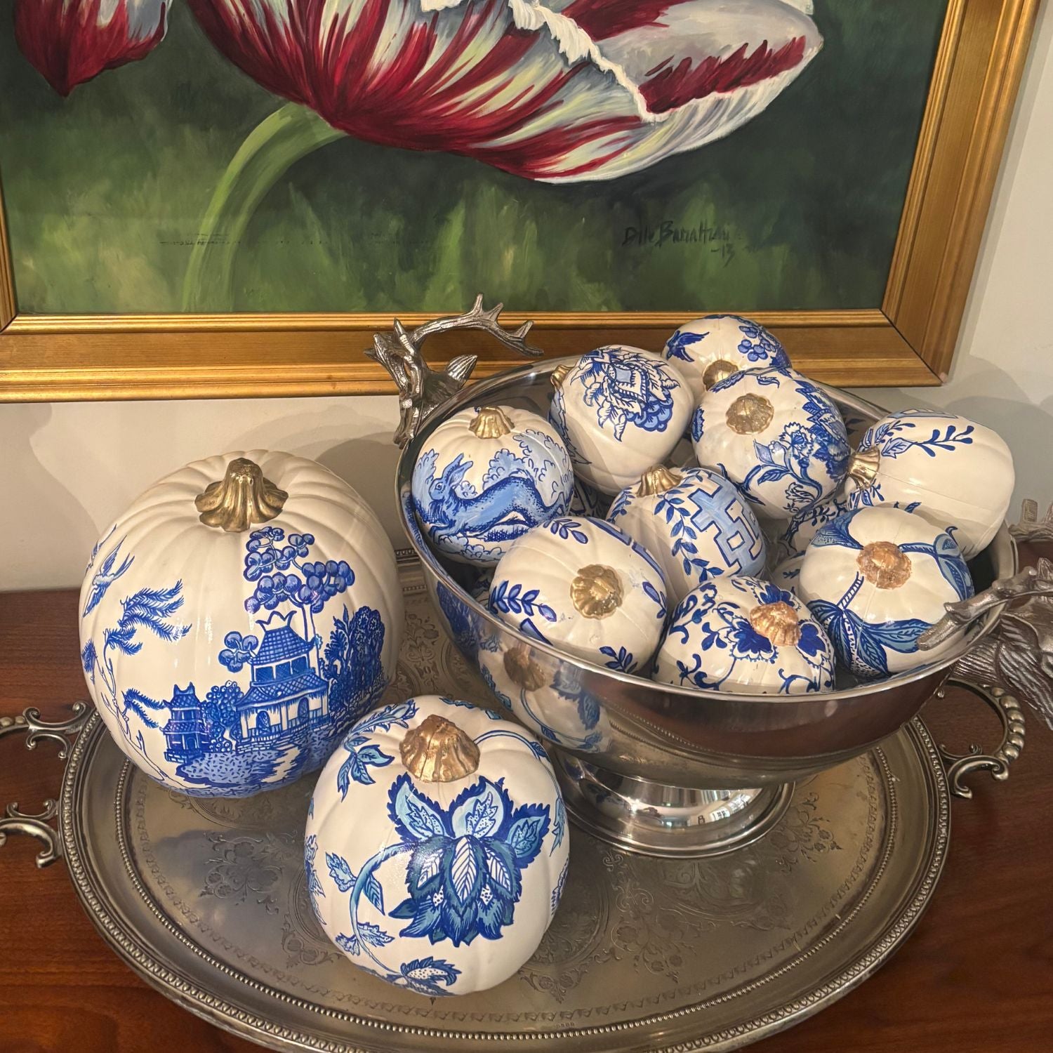 Collection of hand-painted blue and white pumpkins styled in a silver bowl on a wooden sideboard with framed art in the background.