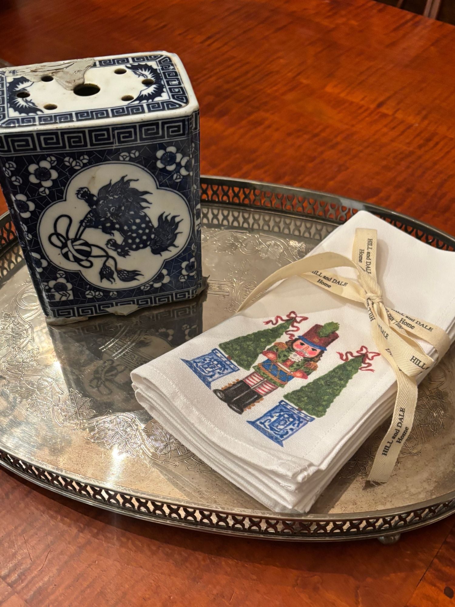 White cloth napkins featuring a festive nutcracker soldier flanked by two green topiary trees in blue and white planters; tied with a Hill & Dale Home ribbon and resting on a silver tray.