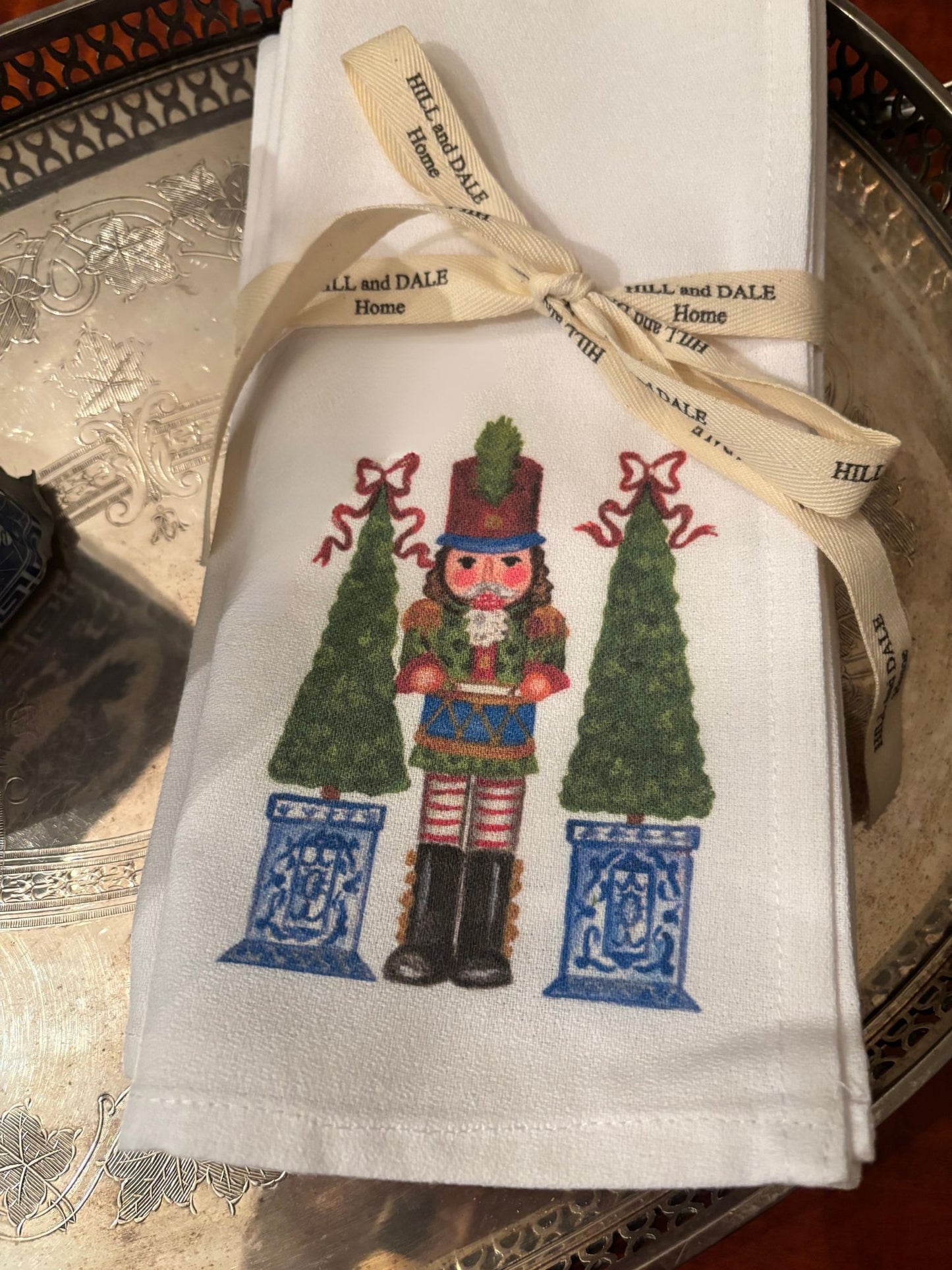 Close up photo of white cloth napkins featuring a festive nutcracker soldier flanked by two green topiary trees in blue and white planters; tied with a Hill & Dale Home ribbon and resting on a silver tray.