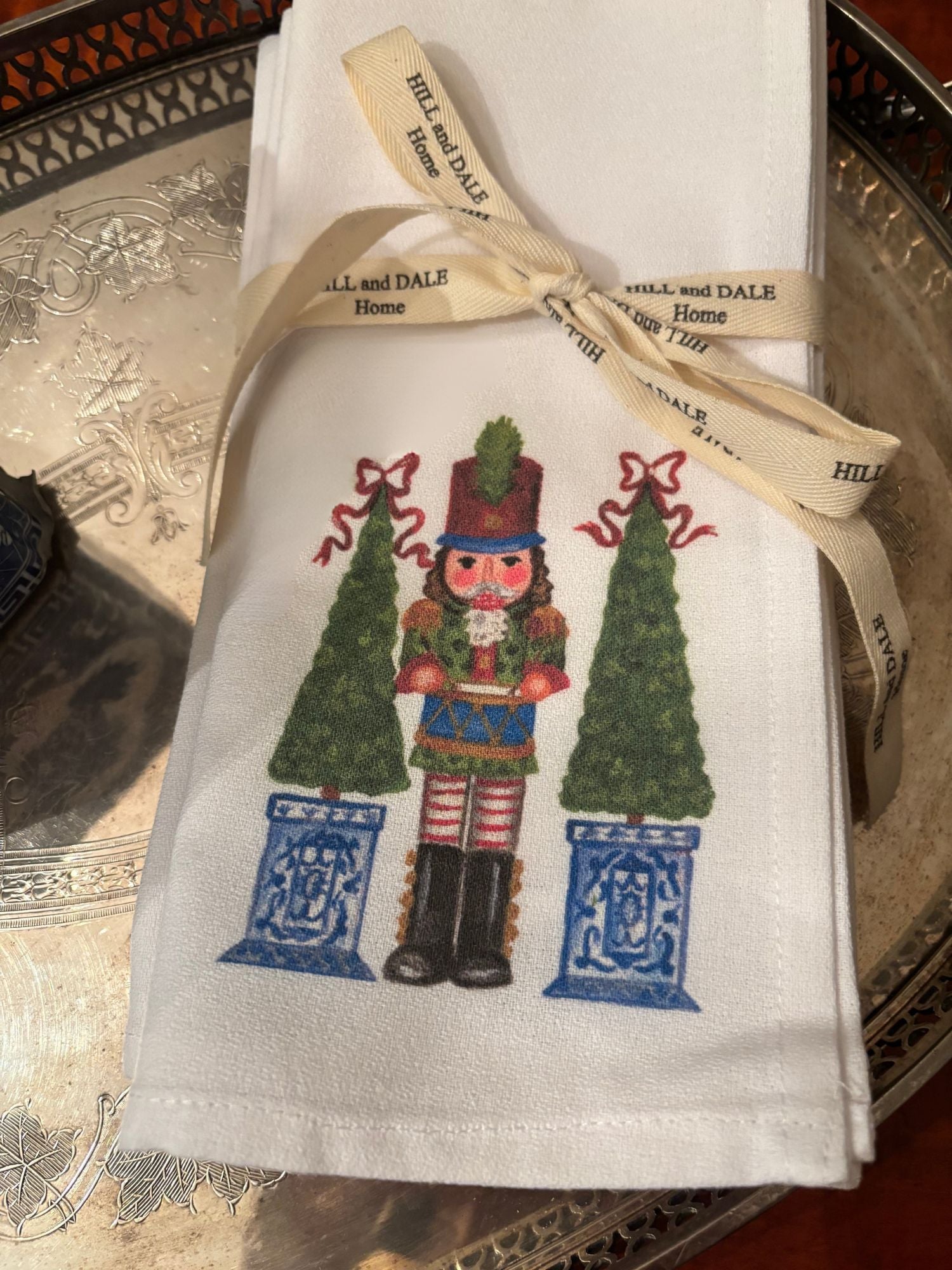 Close up photo of white cloth napkins featuring a festive nutcracker soldier flanked by two green topiary trees in blue and white planters; tied with a Hill & Dale Home ribbon and resting on a silver tray.