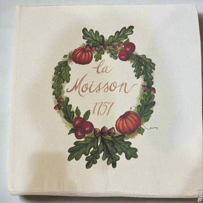 Photo of a square beige throw pillow cover with a wreath of fall harvest designs featuring pumpkins acorns, cranberries and green oak leaves with the words "la Moisson 1757" in the center. 