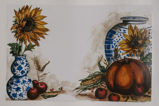 Photo of a paper placemat against a white background. The placemat features a fall harvest of ruby red apples, corn, and a pumpkin next to delicate chinoiserie vases and sunflowers.