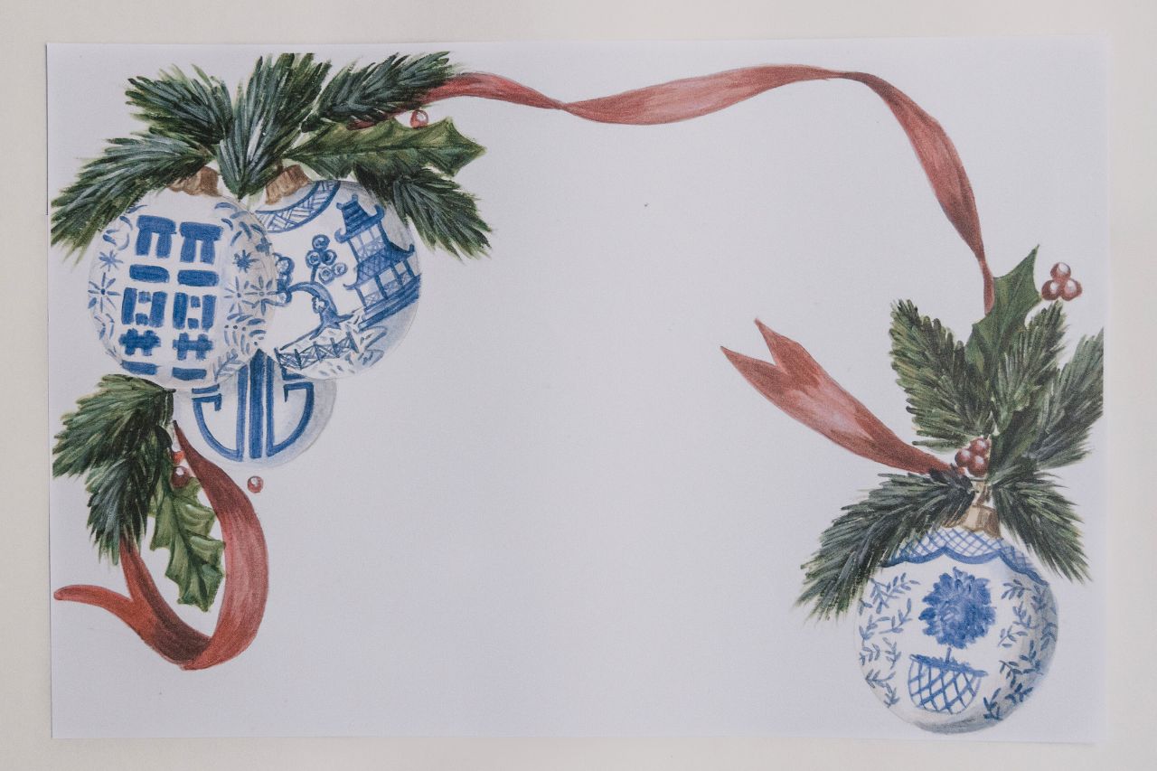 Photo of a paper placemat against a beige background. The placemat artwork features blue and white chinoiserie ornaments, festive greenery, and a bold red ribbon design