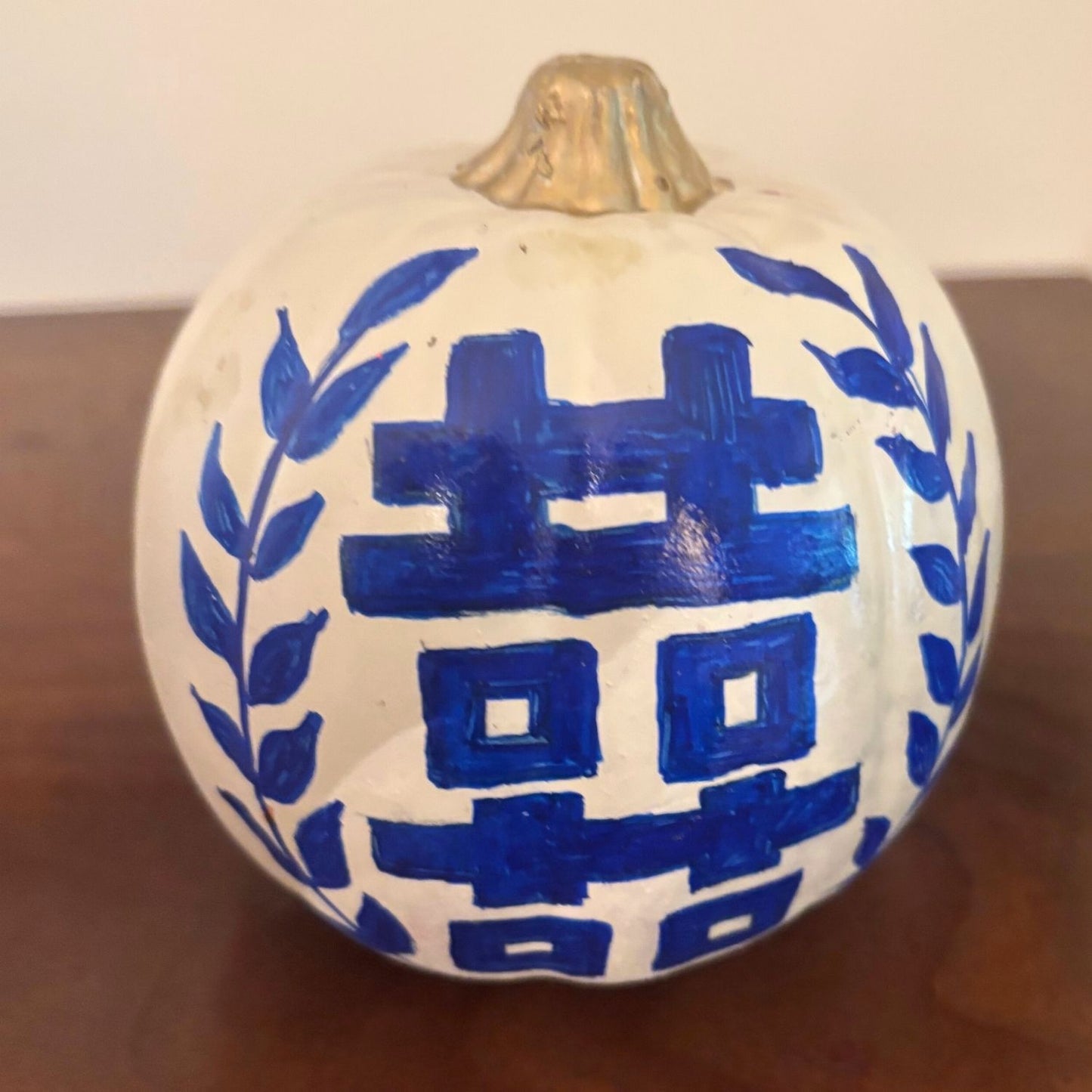 Back side of a hand-painted pumpkin featuring a blue double happiness symbol flanked by laurel branches and topped with a metallic gold stem.