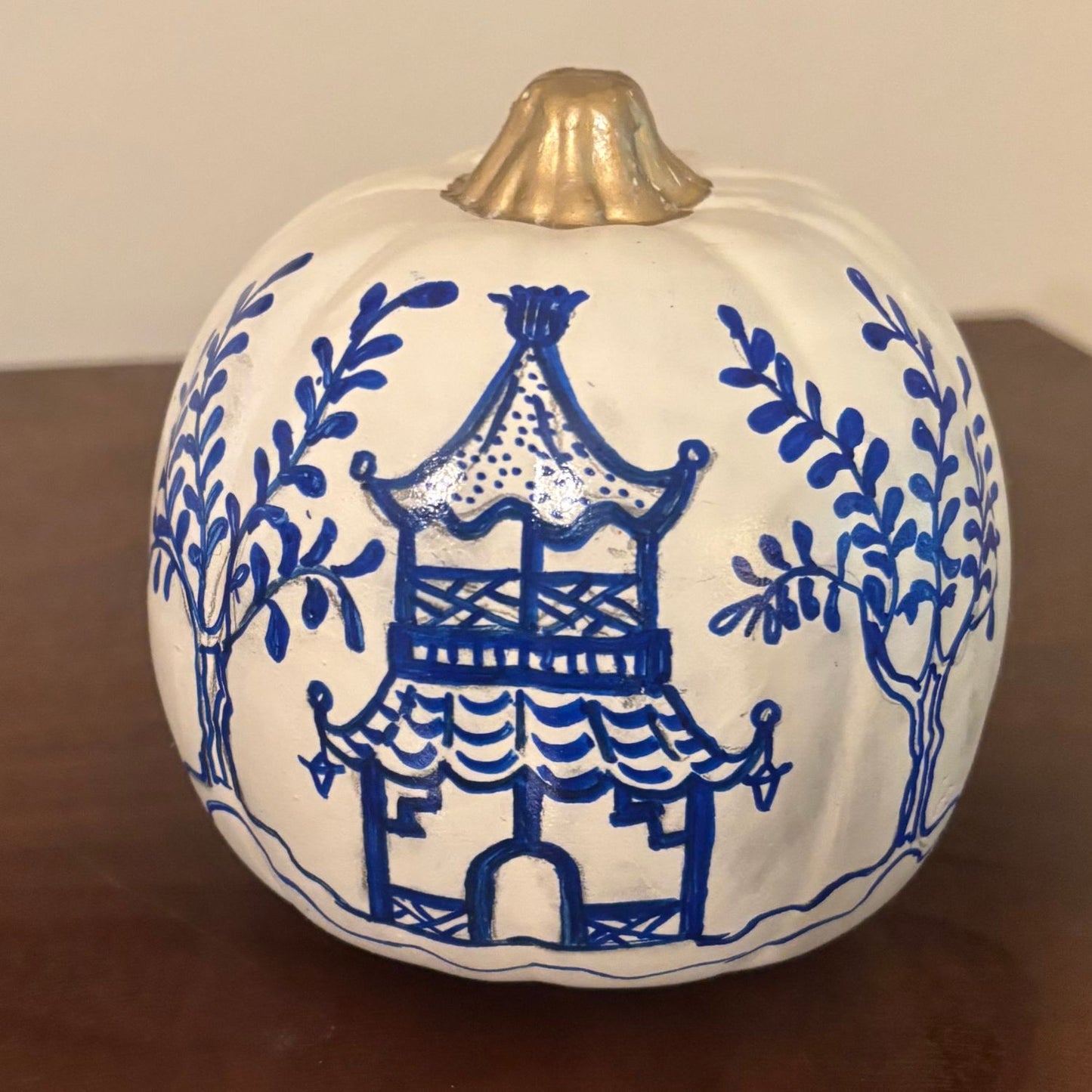 Front side of a hand-painted pumpkin, showing a blue Chinoiserie-style pagoda surrounded by leafy trees, with a gold-painted stem.