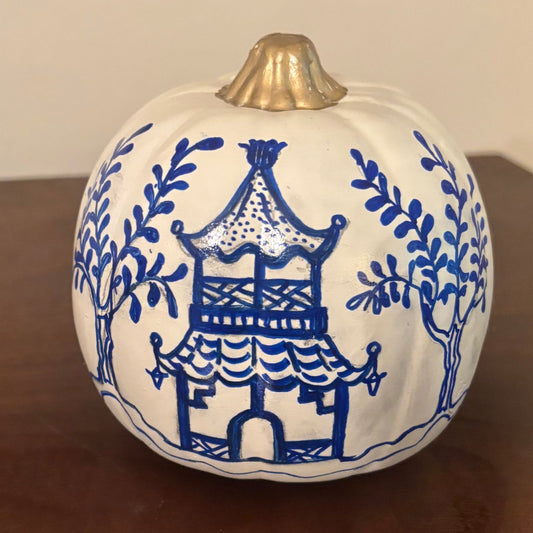 Front side of a hand-painted pumpkin, showing a blue Chinoiserie-style pagoda surrounded by leafy trees, with a gold-painted stem.