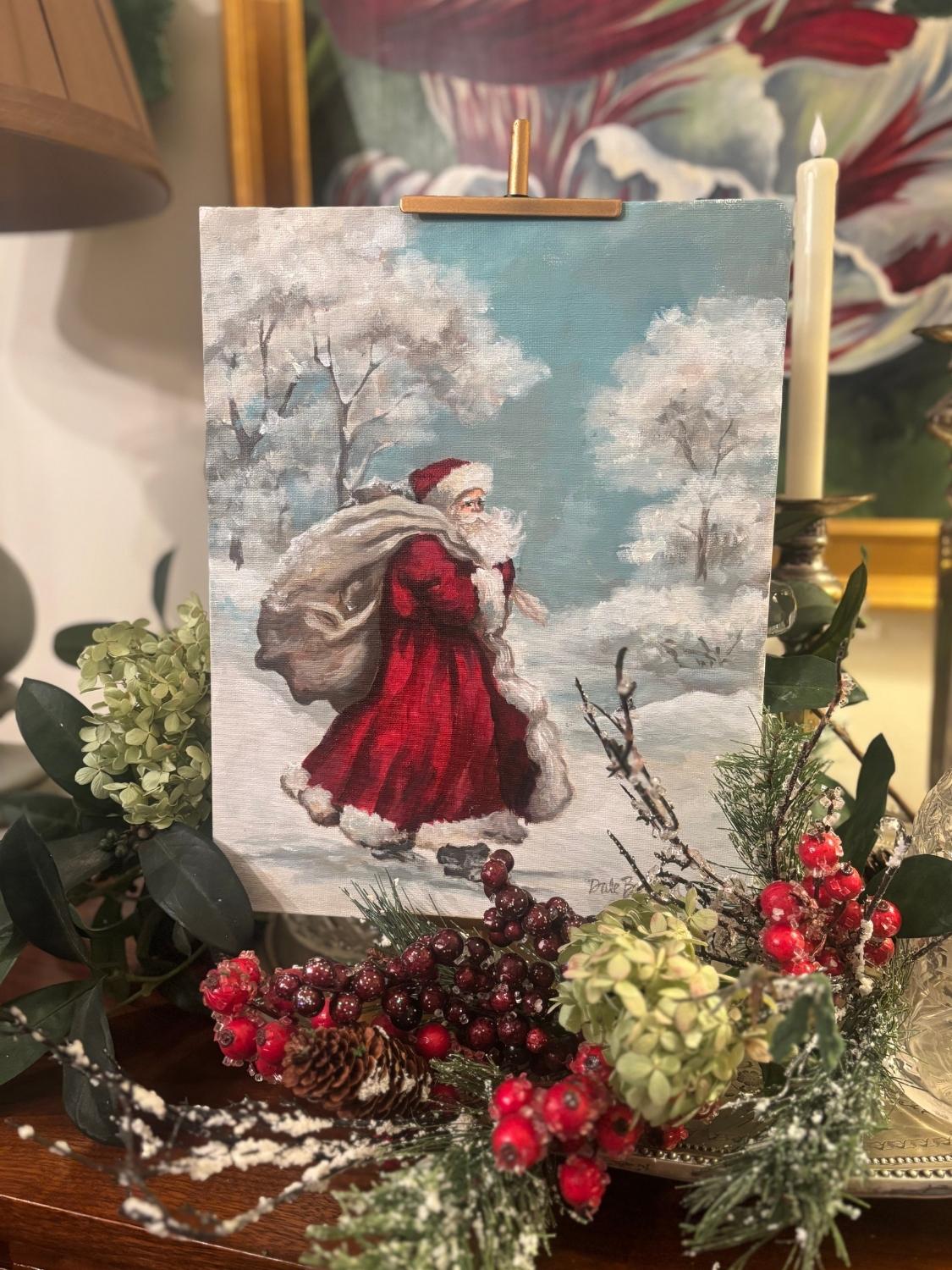 Tabletop art print featuring Santa Claus in a red coat carrying a gift sack as he walks through a snowy woodland scene with frosted trees and a bunny in the background. The art is on a table with greenery and pinecones, decorated for Christmas.
