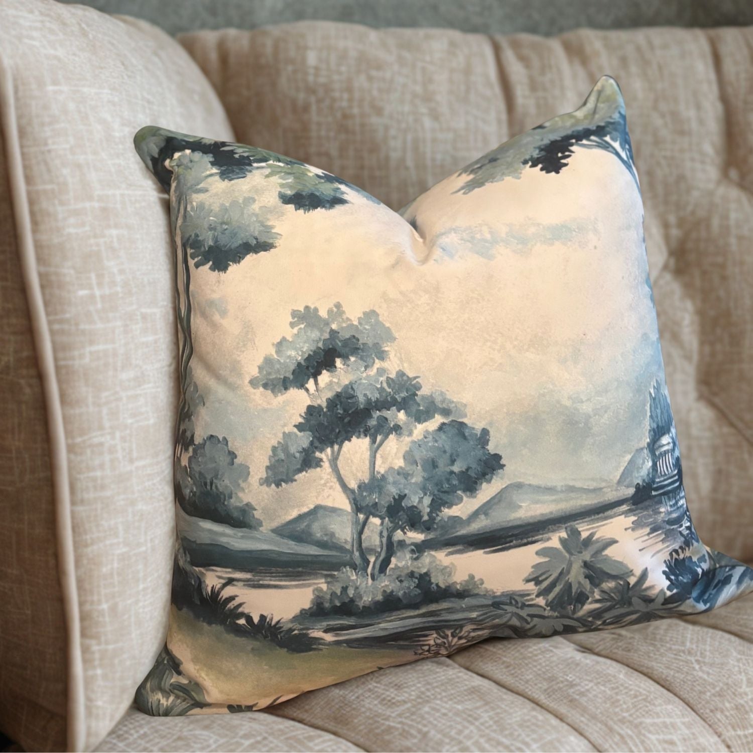 Scenic throw pillow with trees and a river in soft blue and green tones, styled on a beige couch.