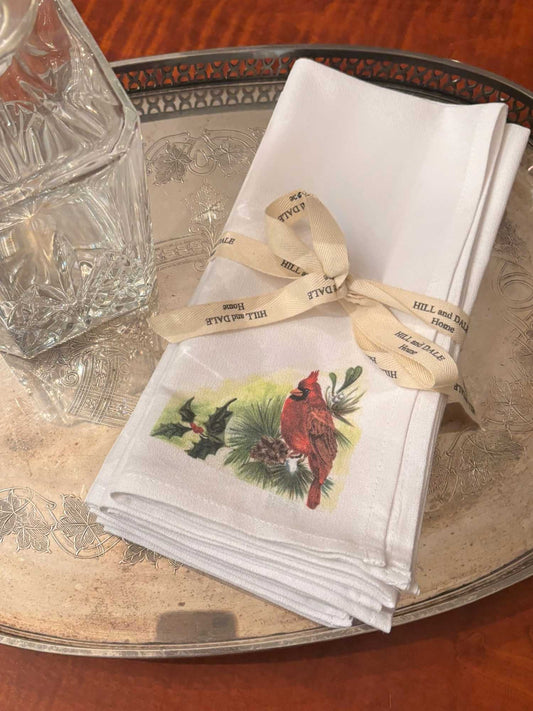 A bundle of four white cloth napkins featuring a red cardinal with holly, a pinecone, and pine sprigs, tied with a Hill & Dale Home ribbon and set on a vintage silver tray.