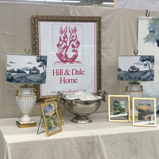 Hill & Dale Home market booth display featuring two elegant white and gold urn-style lamps with hand-painted drum lampshades in a monochromatic blue pastoral landscape design. Between the lamps is a silver punch bowl, and in front are framed miniature landscape paintings. The booth is framed by a large gold-framed sign with two rabbits and the Hill & Dale Home logo, set against a neutral linen backdrop.