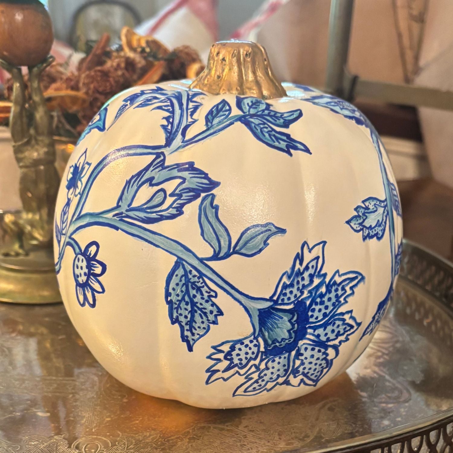 Hand-painted white pumpkin featuring intricate blue floral and leafy vine designs with a metallic gold stem, styled on a vintage silver tray.