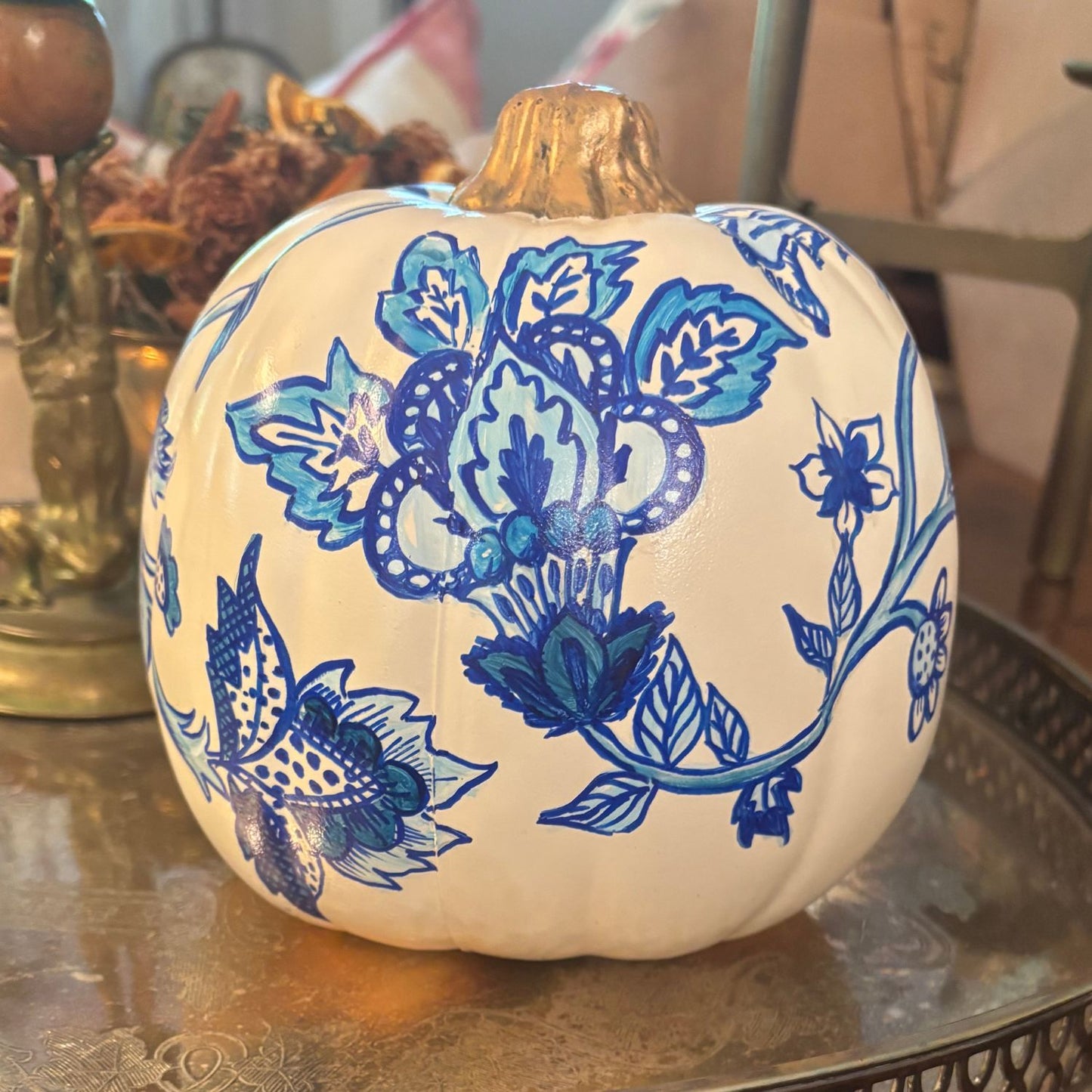 Chinoiserie-style pumpkin adorned with bold blue flowers, trailing vines, and a rich gold-painted stem, displayed on a silvery tray.