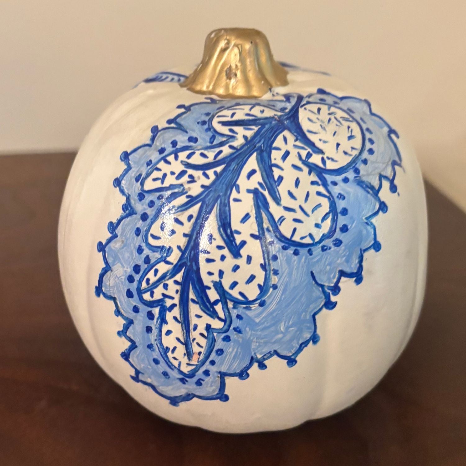Hand-painted white pumpkin featuring a detailed blue oak leaf pattern with dotted accents, topped with a shiny gold stem.