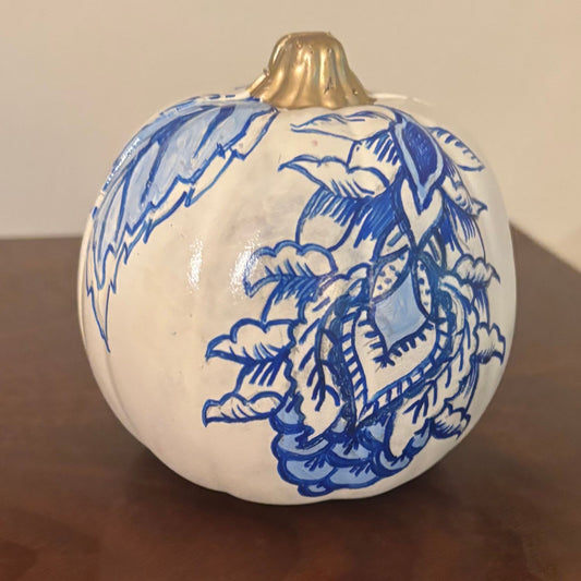 Hand painted white pumpkin with intricate blue leaf and floral design enveloping a conifer, finished with a metallic gold-painted stem.