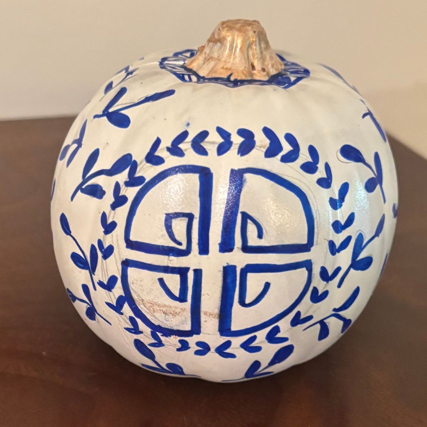 A hand-painted pumpkin featuring a circular prosperity-inspired blue symbol surrounded by delicate vine detailing, on a white base with a gold stem.