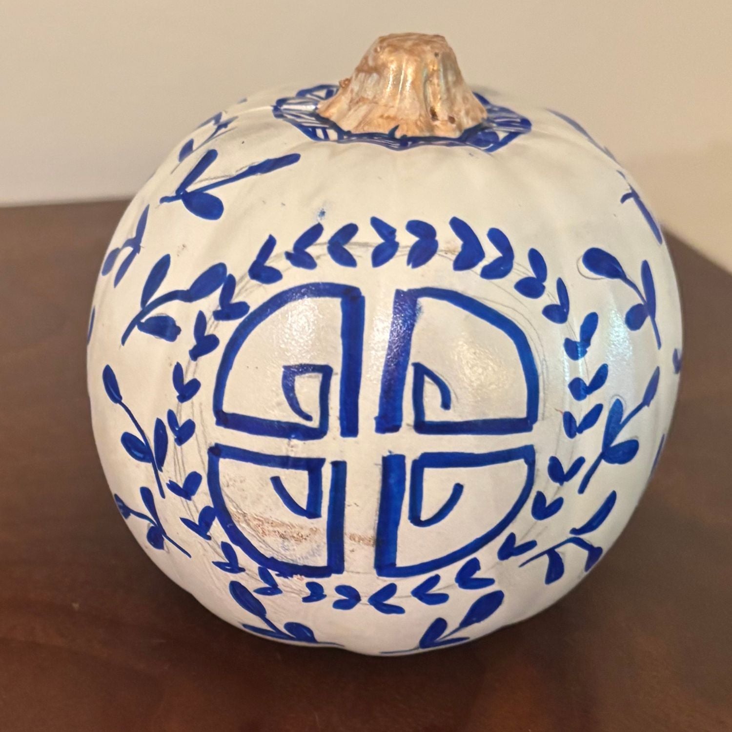 A hand-painted pumpkin featuring a circular prosperity-inspired blue symbol surrounded by delicate vine detailing, on a white base with a gold stem.