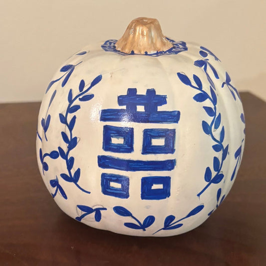 Hand-painted white pumpkin with a bold blue Chinese Double Happiness symbol framed by leafy vines, topped with a gold-painted stem.