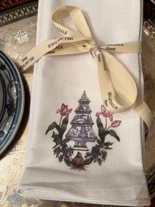 Elegant white cloth napkins with a blue chinoiserie Christmas tree surrounded by red tulips and winter greenery, tied with Hill & Dale Home ribbon.