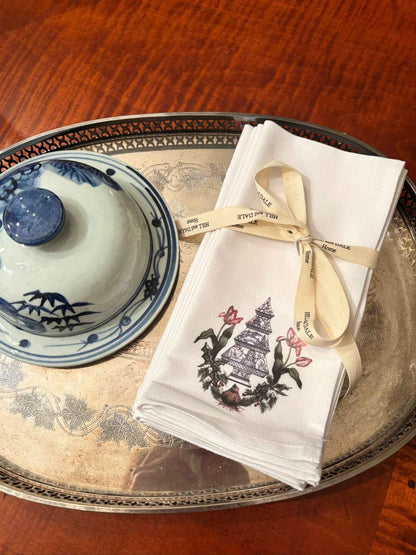 Elegant white cloth napkins with a blue chinoiserie Christmas tree surrounded by red tulips and winter greenery, tied with Hill & Dale Home ribbon and displayed on a silver platter