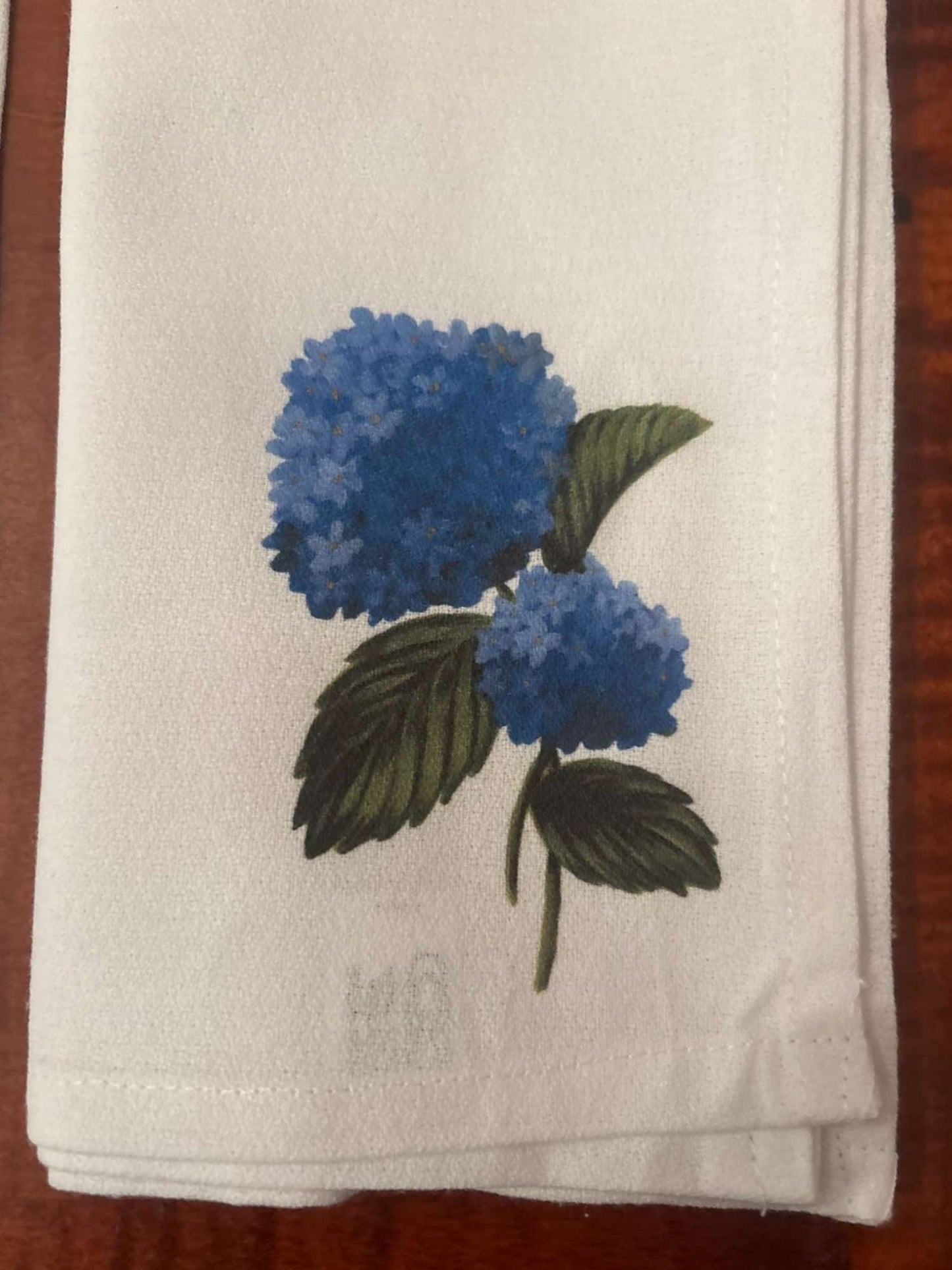 Close view of a white cloth napkin with blue hydrangea design on a wooden surface