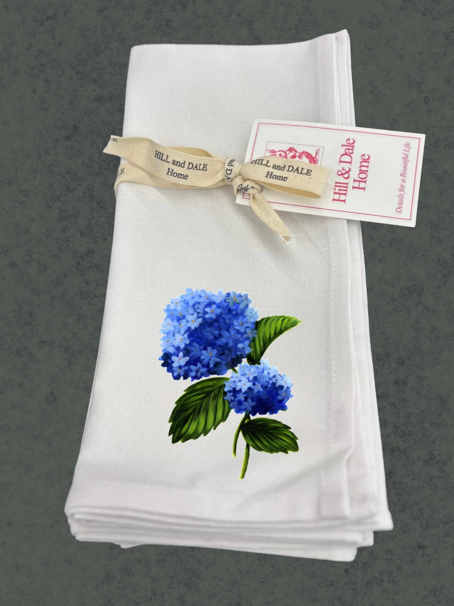 White napkin with blue hydrangea design and 'Hill & Dale Home' tag on a gray background