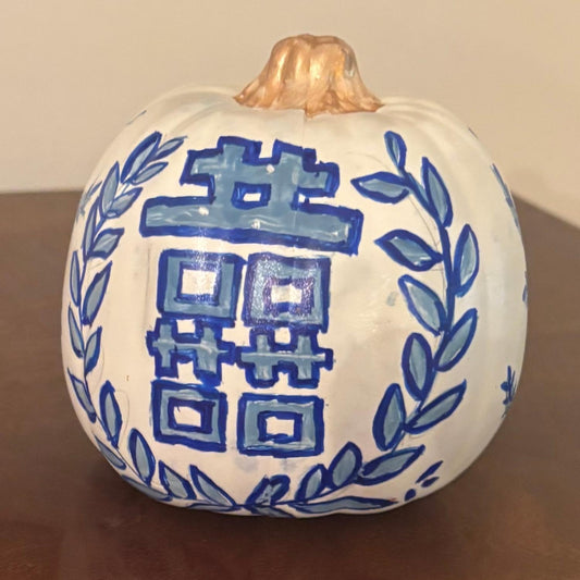 Hand-painted chinoiserie pumpkin featuring a bold blue Double Happiness symbol framed by laurel leaves, with a golden stem on a wooden surface.