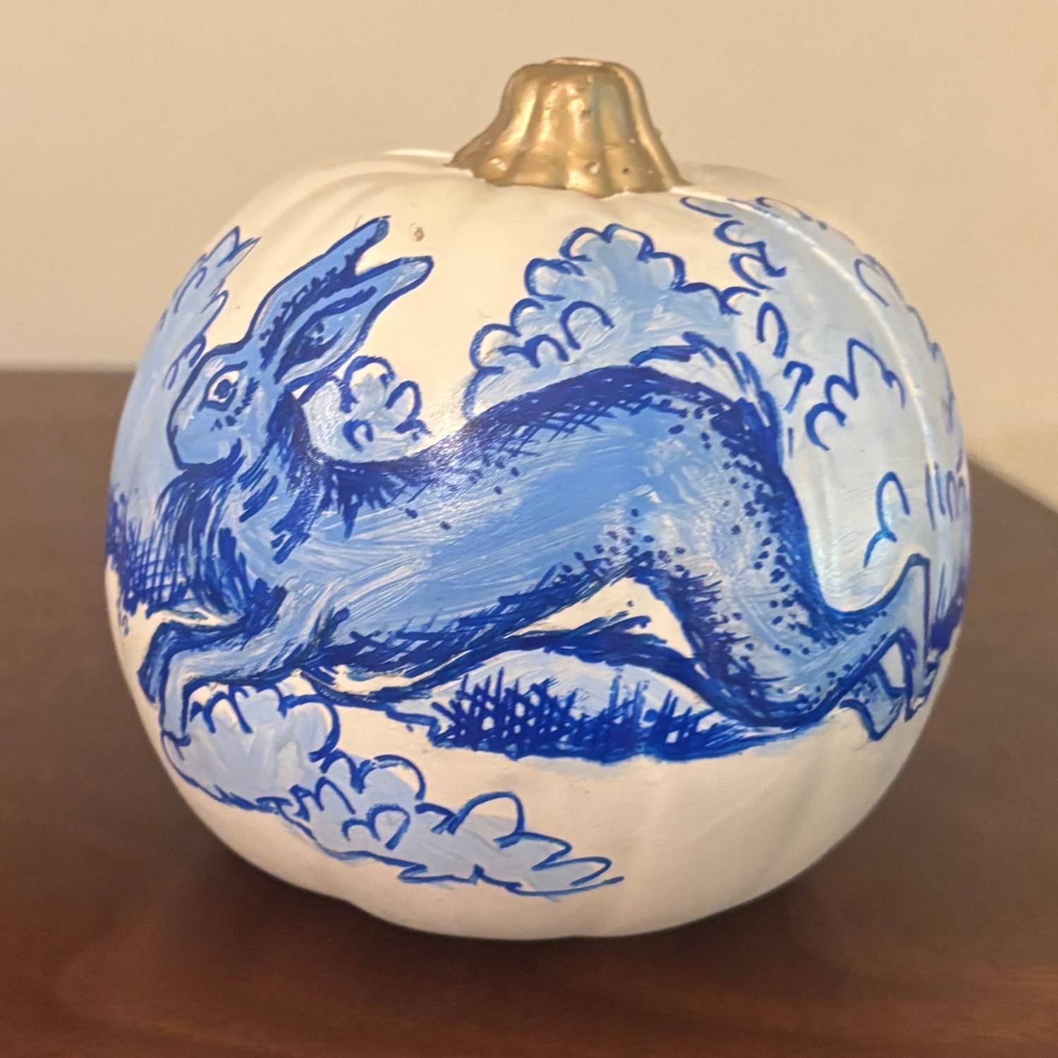 A white decorative pumpkin with a glossy gold stem, hand-painted in blue with a whimsical rabbit leaping through swirling clouds and textured brushstrokes, in the style of chinoiserie porcelain. Displayed on a wooden tabletop with a neutral background.