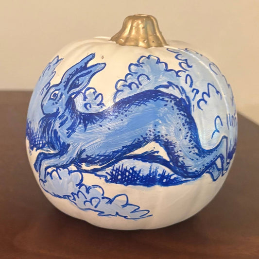 A white decorative pumpkin with a glossy gold stem, hand-painted in blue with a whimsical rabbit leaping through swirling clouds and textured brushstrokes, in the style of chinoiserie porcelain. Displayed on a wooden tabletop with a neutral background.
