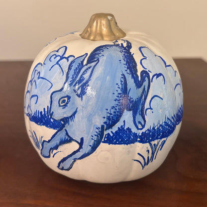 A decorative hand-painted white pumpkin, featuring a blue rabbit in motion with stylized clouds surrounding it. The pumpkin maintains the chinoiserie-inspired aesthetic with rich blue detailing and a metallic gold stem, resting on a wooden surface.