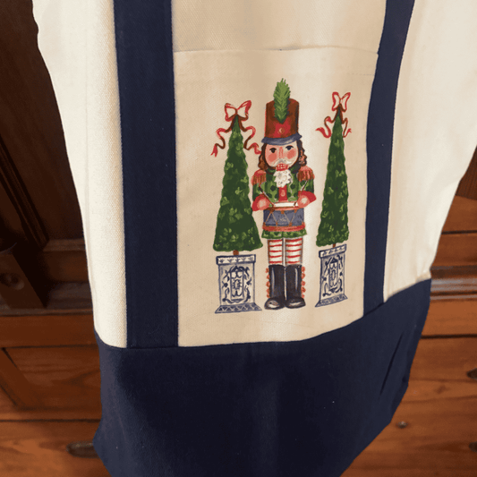 Canvas tote bag with hand-painted nutcracker design, flanked by two holiday topiaries in blue chinoiserie planters