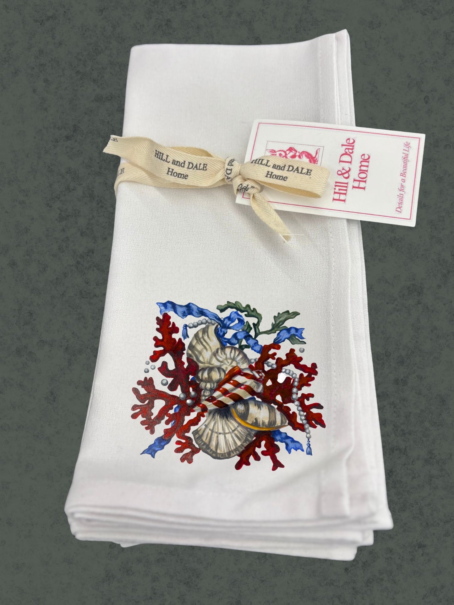 Set of white Hill & Dale Home cloth napkins tied with branded ribbon, featuring Dale Barattini McLin artwork of red coral, seashells, pearls, and blue ribbon accents.