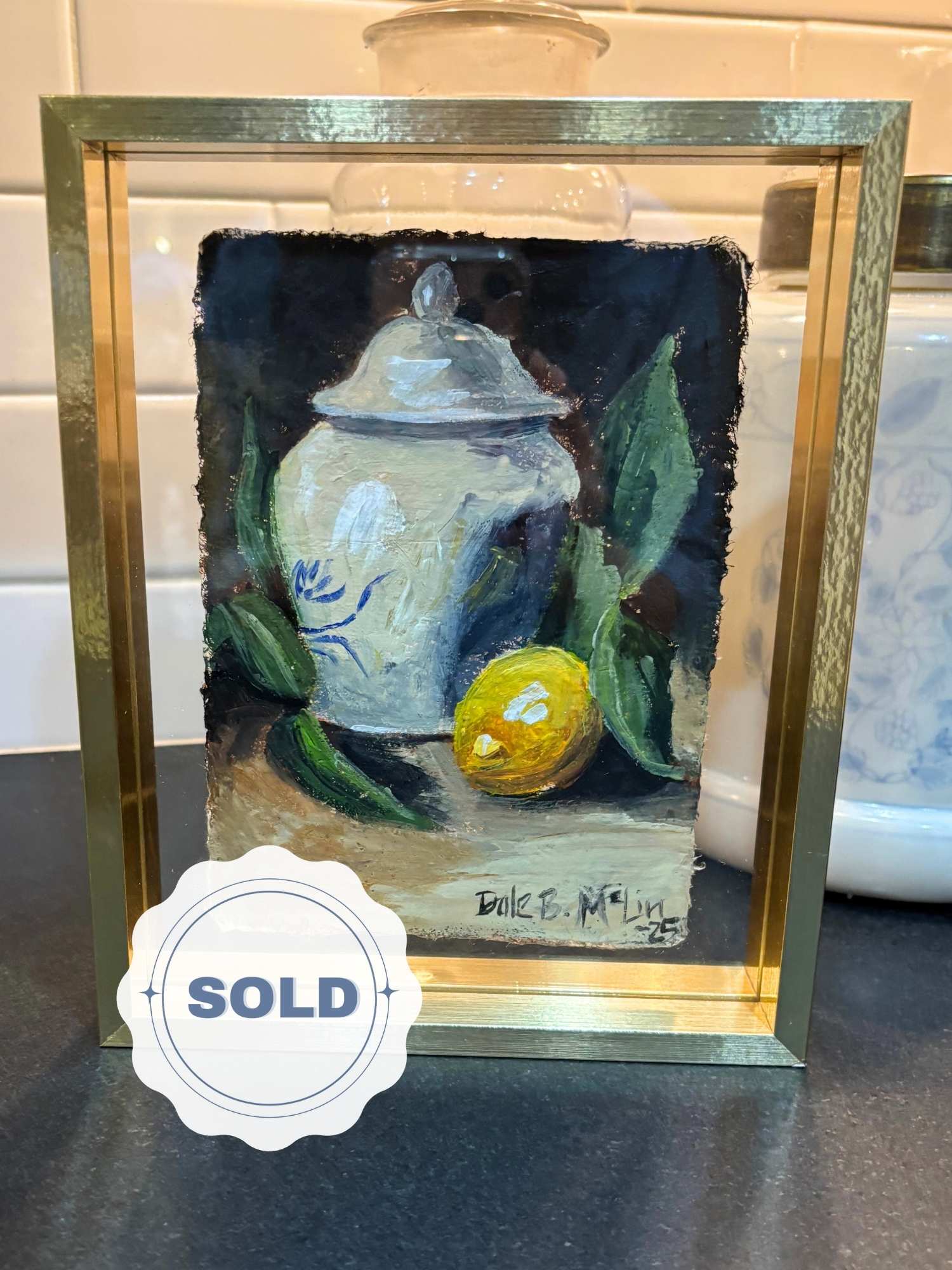 Original acrylic still life of a lemon beside a blue and white porcelain jar, painted on handmade Lokta paper and framed in a brass floating frame. Sold icon layered over product photo in bottom left corner. 