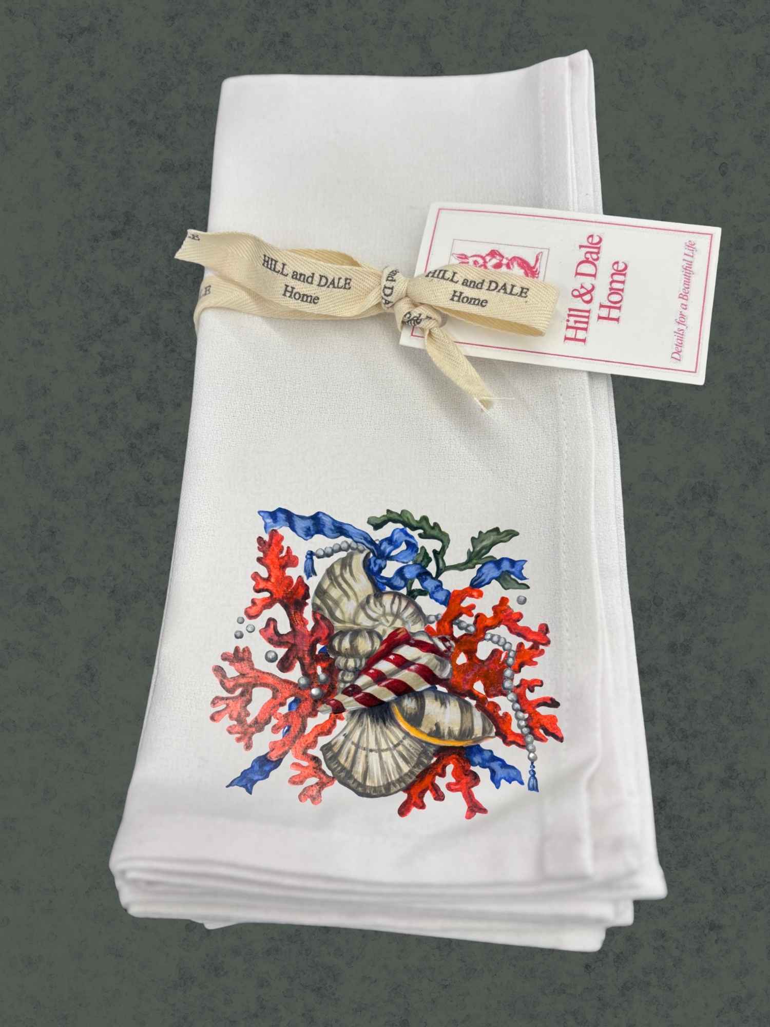 Set of white Hill & Dale Home cloth napkins tied with branded ribbon, featuring Dale Barattini McLin artwork of sunset coral, seashells, pearls, and blue ribbon accents.